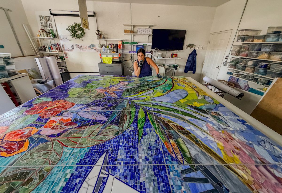 Mila Apperlo, CEO of Apperlo Art, sits behind a monumental handcrafted glass mosaic in the studio, surrounded by neatly organized shelves of materials and tools. The vibrant artwork depicts lush tropical leaves, blooming flowers, and flowing blue water, showcasing the scale, artistry, and craftsmanship behind the creative process.